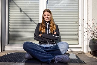 Junges Mädchen in Arbeitskleidung sitzt vor einem Rollladen und lächelt in die Kamera